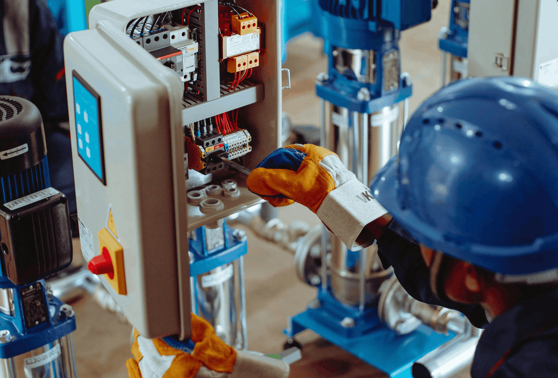 Technician in a hard hat and gloves working on the wiring inside an open control panel beside commercial pumps